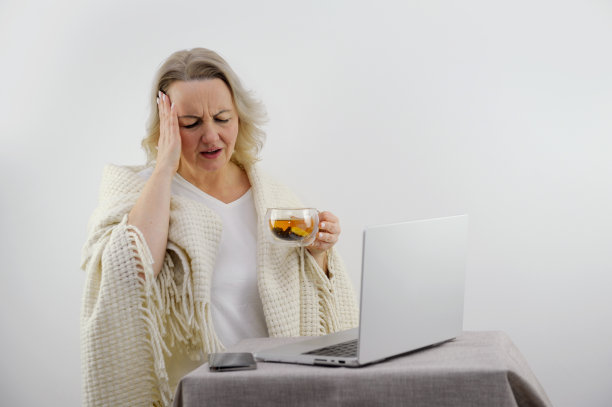 我的黑发女孩坐在沙发上，用笔记本电脑远程工作。女在擤鼻涕，发烧和头痛。年轻女子在家治疗，请了病假。冬季感冒和流感的概念图片下载