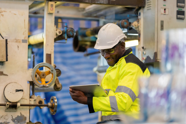 非洲男工程师当机械师的特写照片，拿着笔记本电脑检查工作，在塑料和钢铁行业工作，微笑着，戴着安全帽，穿着背心。有一个电子电路在工作。图片下载