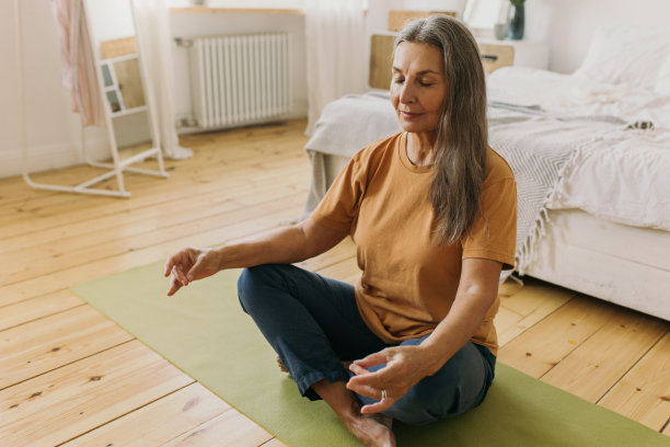 室内图片:运动的成熟女性用手做手印，深呼吸，冥想，闭上眼睛，坐在绿色垫子的地板上，以卧室为背景，有床，窗户和镜子图片下载