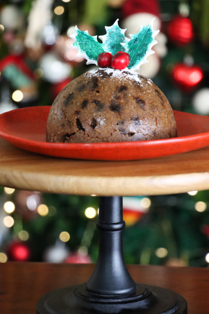 特写图片:自制的红盘子，蛋糕架上装饰着冬青叶和红浆果的圣诞布丁，喜庆的圣诞树和照亮的仙女灯背景，重点放在前景上图片下载