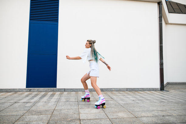 在白色的墙壁背景下，年轻的女人用五颜六色的辫子在外面的复古滚轴上溜冰图片下载