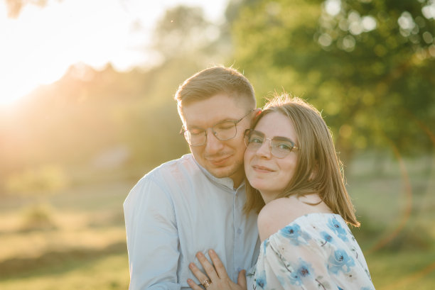 在夕阳的背景下，一对幸福的情侣在田野上拥抱。夏日公园里的男女肖像。夕阳下，美丽的女人抱着戴眼镜的男人站在绿草地上。特写照片。图片下载