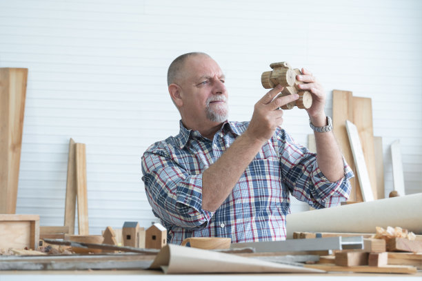 在车间工作的高加索老白胡子木匠，拿着，细细地看着手工木制汽车玩具，工具机器和小的木制模型或手工艺品在白色房间的桌子上图片下载
