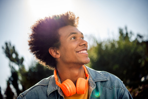 照片上的年轻人笑容满面，穿着牛仔裤夹克，脖子上戴着耳机，在公园里享受阳光明媚的天气图片下载