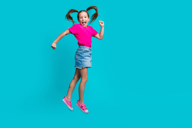 全身照片喜出望外的飞行女孩穿着时尚的t恤牛仔裙跳跃握紧拳头孤立在蓝绿色的背景图片下载