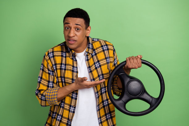 照片中的年轻人穿着黄色的格子衬衫，展示大众汽车的方向盘孤立在绿色的背景上图片下载