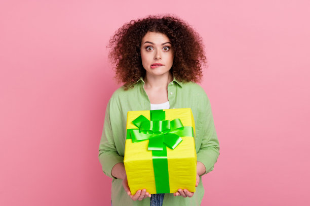 漂亮的年轻女孩的照片肖像拿着紧张的礼盒咬嘴唇担心穿着时髦的绿色服装孤立在粉红色的背景图片下载