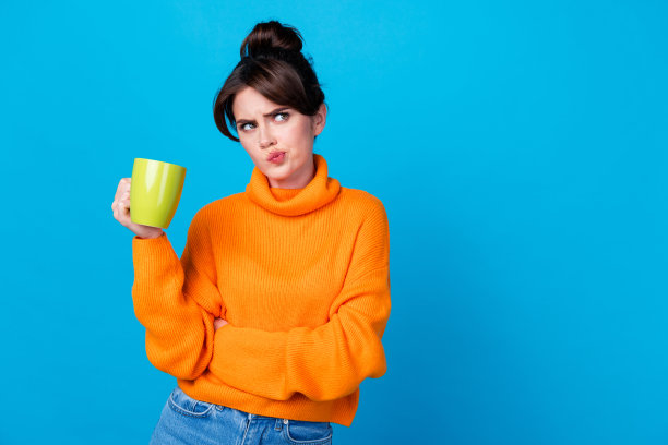 照片中疑神疑神的女人穿着橙色毛衣喝咖啡，看起来空无一物，孤立的蓝色背景图片下载