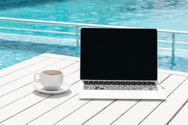 在度假酒店泳池背景的桌子上放着黑屏笔记本电脑和咖啡杯。工作与休闲结合，在线远程数字化教育，远程学习，另类办公图片下载