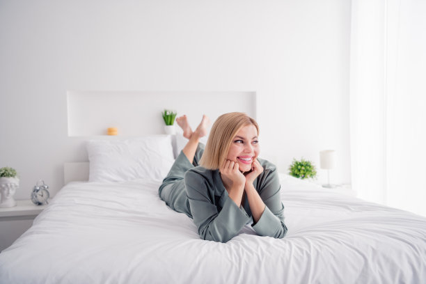 年轻躺在床上的照片，漂亮的微笑，可爱的女人抚摸脸颊，看着卧室窗户里的阳光，放松地穿着睡衣图片下载