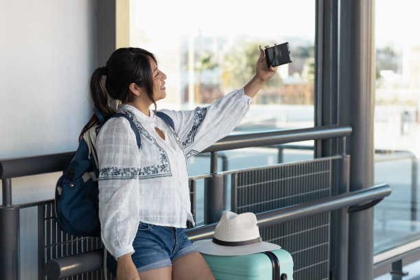 一名26岁的女子，带着行李箱和背包到达墨西哥的目的地，在交通车站用手机拍了一张照片。图片下载