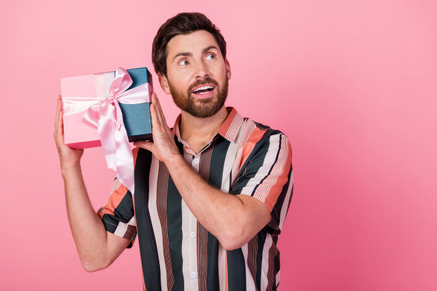 照片英俊的成熟男子与胡茬看起来新奇买了可爱的包装礼物生日快乐孤立的粉红色背景图片下载