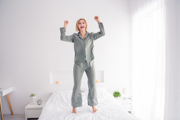 照片肖像金发迷人的年轻女子跳床有乐趣穿着时尚的灰色睡衣轻现代卧室极简主义的设计图片下载