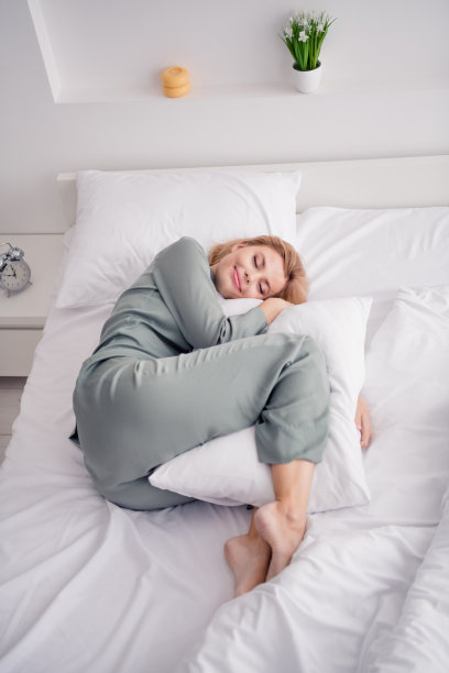 照片肖像金发迷人的年轻女子躺在床上抱枕头穿时髦的灰色睡衣灯卧室室内极简主义的设计图片下载