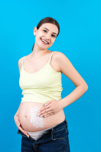 一名孕妇穿着拉开拉链的牛仔裤，在腹部涂上润肤霜，衬托出彩色背景上的妊娠纹。护肤理念图片下载