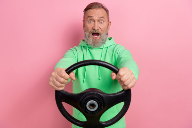 照片中，激动的印象深刻的男子穿着绿色运动衫骑着快速的汽车，孤立的粉红色背景图片下载