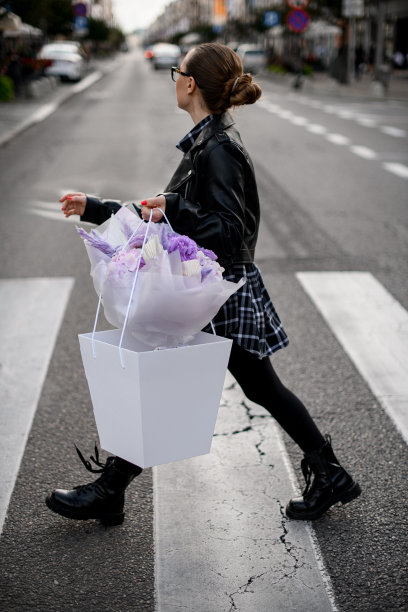 可爱的年轻女子正在人行横道上过马路，手里拿着一大束紫色的花、玫瑰、绣球花和其他装饰元素，装在礼品盒袋里图片下载