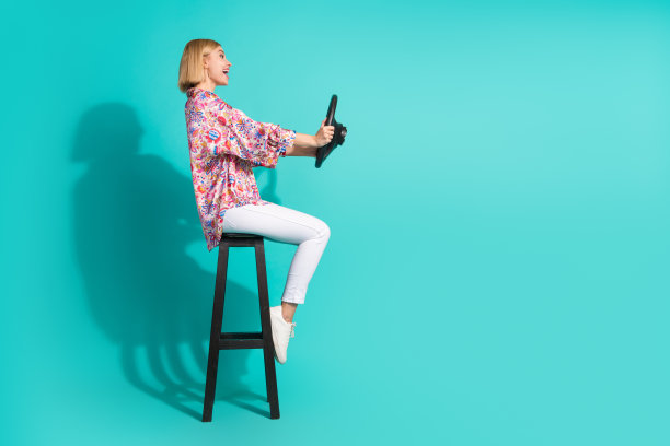 漂亮年轻女孩的全身照片，坐在椅子上，握着方向盘，穿着时髦的粉红色印花服装，孤立在青色背景上图片下载