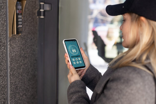 女性手持黑色智能手机，屏幕上有智能家居应用。背景上模糊的房子。为了进入你的房子的所有控制和家庭安全的关怀图片下载