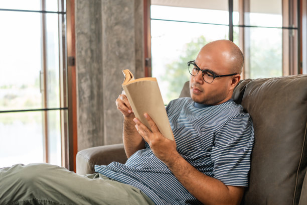 亚洲人坐在舒适的沙发上看书在客厅与阳光的早晨。人们有时呆在家里放松一下。美好时光品质生活安宁幸福图片下载