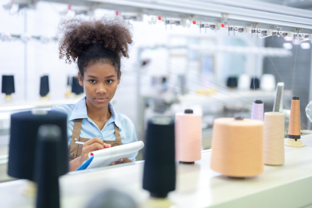 美国女设计师，在一家工业工厂里，站在等待发给顾客的服装和织物生产机器旁边，看着线轴，写着一张纸条。图片下载