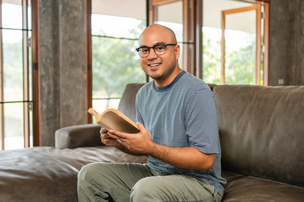 亚洲人坐在舒适的沙发上看书在客厅与阳光的早晨。人们有时呆在家里放松一下。美好时光品质生活安宁幸福图片下载