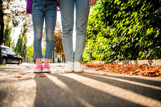 裁剪的照片两个女孩的腿牛仔牛仔裤运动鞋阴影道路落叶绿色公园外的灌木丛图片下载