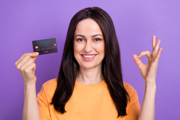 照片中，一个长着牙齿、笑容灿烂的聪明女人穿着橙色的t恤，手里拿着一张信用卡，上面写着“ok”，建议在紫色的背景上孤立起来图片下载