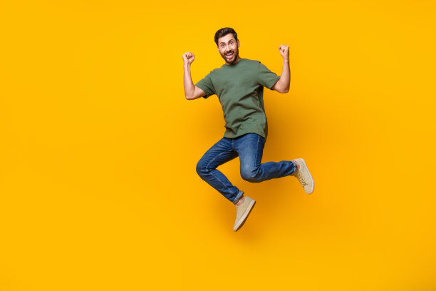 迷人的年轻人的全身照片跳跃，举起拳头庆祝穿着时尚的卡其布衣服孤立的黄色背景图片下载