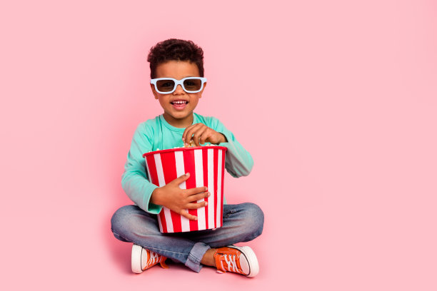 可爱小男孩吃爆米花的全身照片3d眼镜坐在地板上穿时髦的海蓝宝石衣服孤立在粉红色的背景图片下载