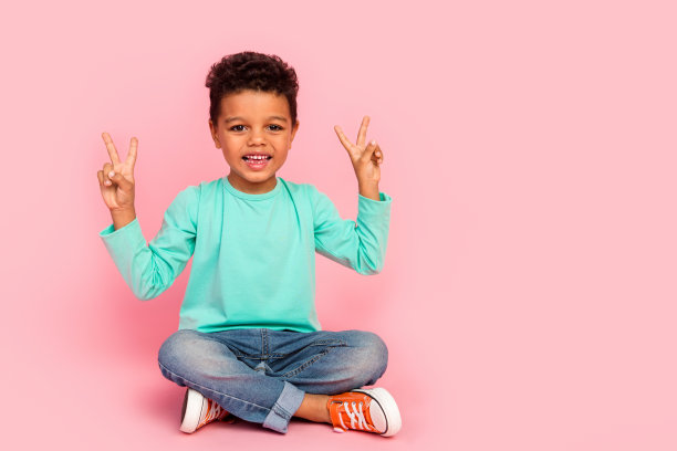 可爱小男孩的全身照片坐在地板上显示双v字穿着时髦的海蓝宝石衣服孤立在粉红色的背景图片下载