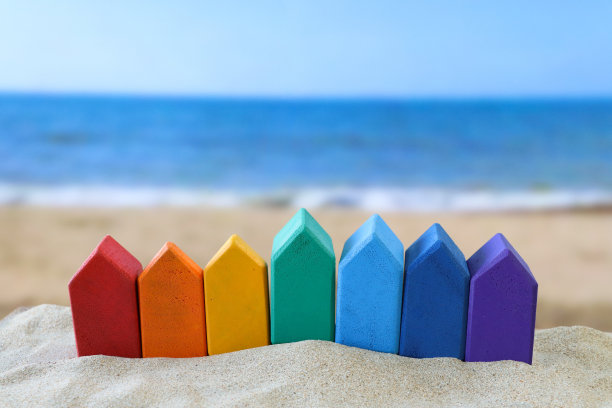 一排彩虹木制模型度假小屋的特写图片，在一堆沙子上，红色，橙色，黄色，绿色，浅蓝和深蓝，紫色，沙滩背景，海浪在退潮时破碎，大海和海岸线，度假租赁和度假屋的概念图片下载