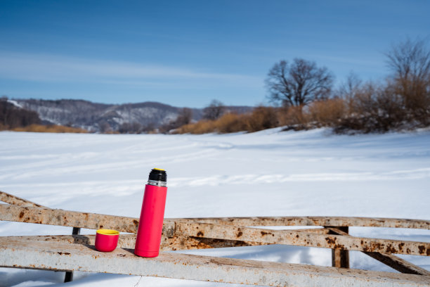 红色的保温瓶放在雪白的木栅栏上，以自然景观为背景图片下载