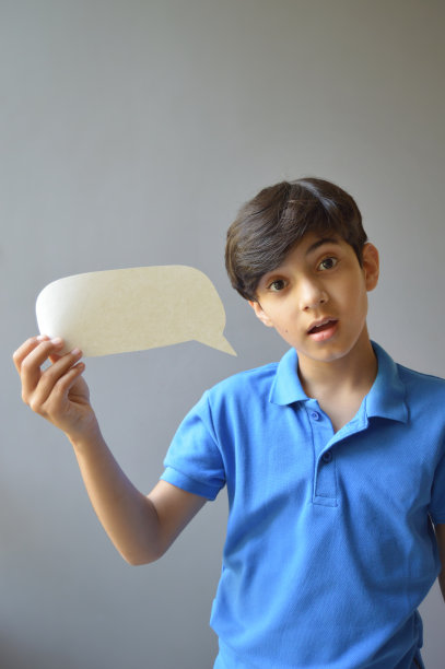 一个可爱的11岁男孩的垂直照片，穿着休闲的蓝色polo t恤，拿着一个纸工艺品，剪纸的形状是一个空白的空洞的演讲泡泡或号召气球，在灰色的背景下，带着惊讶的表情看着镜头图片下载