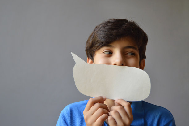 一个可爱的11岁男孩的横向照片，穿着休闲的蓝色t恤，微笑着拿着一个纸工艺品，剪纸成一个空白的空演讲泡泡或号召气球的形状作为面具，看着灰色背景上有趣的微笑，翻白眼图片下载