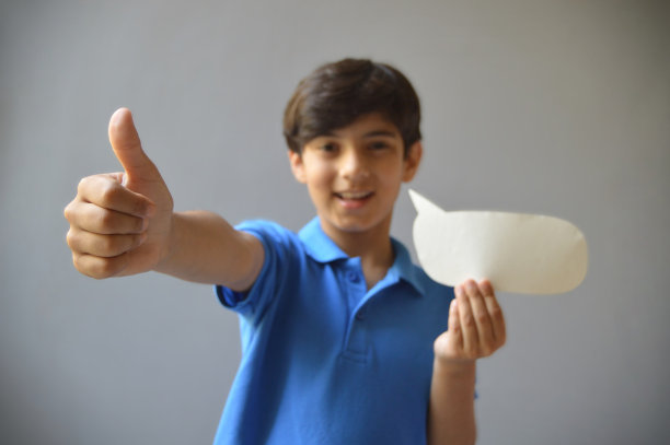 一个可爱的11岁男孩的横向照片，穿着休闲的蓝色polo t恤，手里拿着一个纸工艺品，剪纸的形状是一个空白的空洞的演讲泡泡，或者大声欢呼，或者竖起大拇指的手势，背景是灰色的图片下载