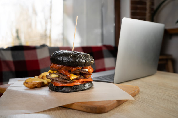 垃圾食品，在咖啡馆或餐馆用笔记本电脑工作时用木板盛着的黑色汉堡和薯条，不健康的生活方式图片下载