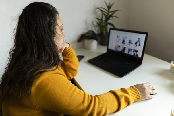 网上购物在家里，美丽的女人使用笔记本电脑在她的家庭工作空间和网上购物。穿着舒适的黄色毛衣。库存图片图片下载