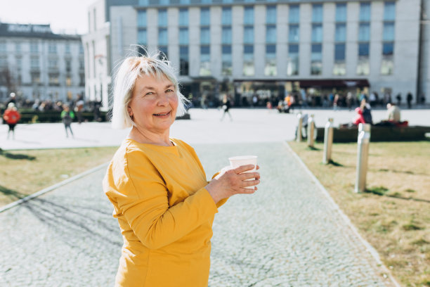 快乐成熟的高加索女人在好心情在城市散步咖啡在白天。生活方式的概念。老人拿着外卖咖啡杯在城市背景下图片下载