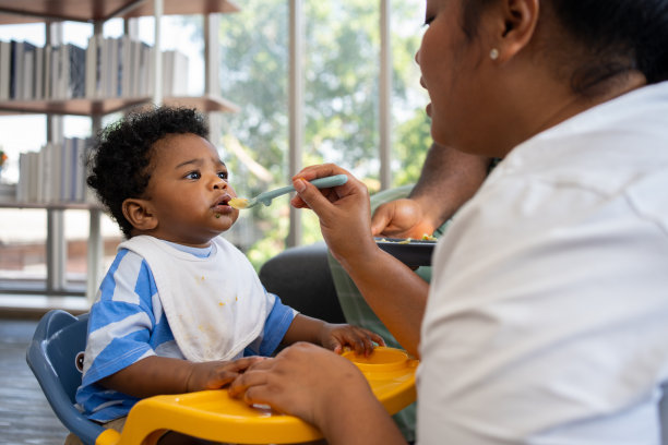 亚洲妈妈在给她9个月大的可爱宝宝喂奶，非裔美国人在家帮忙拿盘子。照片系列的家庭，孩子和快乐的人的概念。父母喂养孩子。图片下载