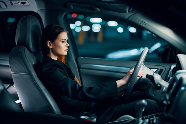 这是一张从侧面拍摄的横向照片，拍摄于夜间，一位身穿黑色衬衫、系着安全带的女性坐在方向盘后面，看着道路。安全驾驶主题图片下载