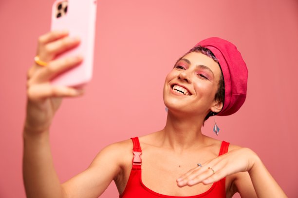 一位留着粉色头发和短发的年轻女博主用手机拍了一张自己的照片，她穿着时髦的衣服，戴着粉红色背景的单色帽子，脸上挂着微笑图片下载