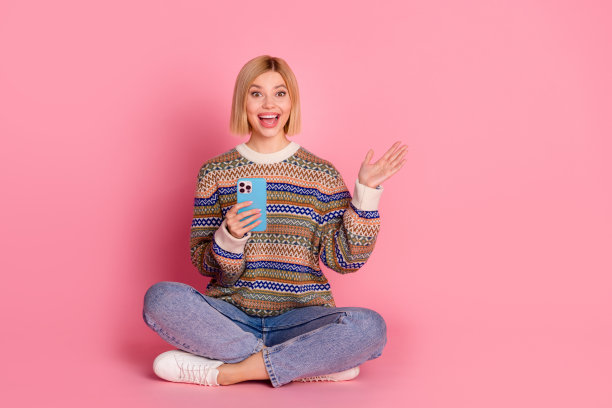 全尺寸照片，印象深刻的女人穿着装饰套头衫，坐在智能手机惊讶地盯着孤立的粉红色背景图片下载
