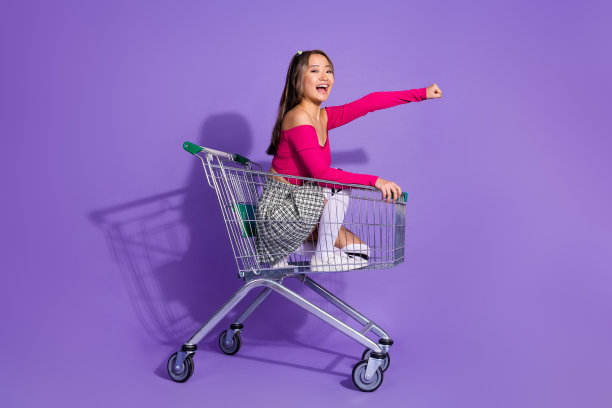 可爱时髦的女士穿着粉色上衣骑着快速超市手推车的全身照片，空旷的空间孤立的紫色背景图片下载