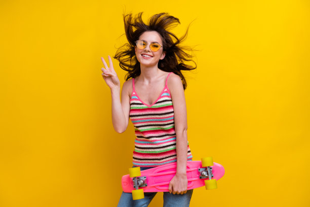 照片中，开朗的好女人穿着针织衫，戴着太阳镜，手持滑板，在充满活力的黄色背景上显示v字图片下载