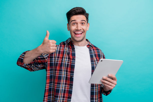 照片中，一个穿着格子衬衫的可爱的兴奋的男人正在和现代电子设备聊天，屏幕上的拇指向上翘起，背景是蓝绿色图片下载