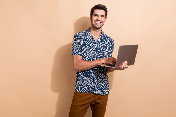 照片聪明、聪明、积极的男人，有时尚的发型，穿着印花衬衫，在笔记本电脑上打字，孤立在米色背景上图片下载