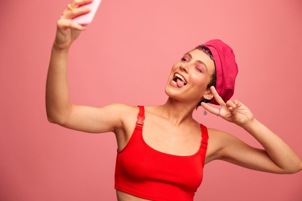 一位留着粉色头发和短发的年轻女博主用手机拍了一张自己的照片，她穿着时髦的衣服，戴着粉红色背景的单色帽子，脸上挂着微笑图片下载