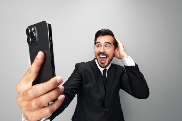 男子商人手里拿着手机惊讶地看着屏幕上的视频通话和自拍，笑得龇牙咧嘴。特写广角照片灰色孤立背景图片下载