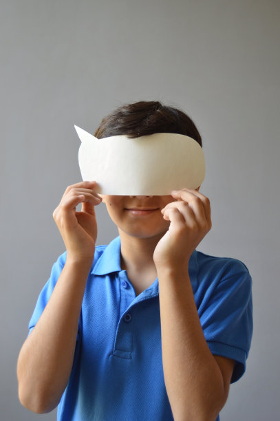 一个穿着休闲蓝色t恤的可爱的11岁男孩的垂直照片，他拿着一个纸剪成一个空白的空演讲泡泡或号召气球的形状，作为面具蒙住他的眼睛。图片下载